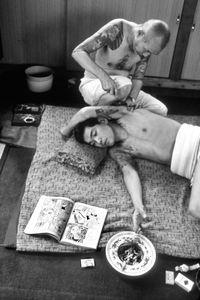 Man reads comic book while getting tattoo by Horibun 1 in Tokyo studio,1970 © Martha Cooper